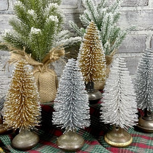 May include: A collection of miniature Christmas trees in various colors, including green, gold, and silver. Some trees are flocked with white, and all are mounted on vintage-style bases. The trees are arranged on a plaid surface.