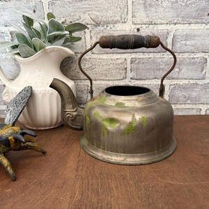 May include: An aged metal kettle with a wooden handle and spout. The kettle shows signs of wear with green staining. A white jug with foliage and a decorative bee are in the background.