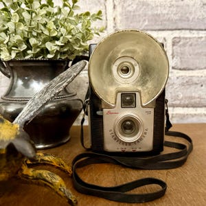 May include: A vintage black Brownie Starflash camera with a large silver circular flash reflector, a silver front faceplate, a Dakon lens, and a black neck strap. The camera body features the text Brownie Starflash Camera 13 Color B&W 14.