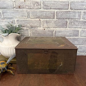 May include: A rectangular, antique metal storage box with a hinged lid and a latch. The box has a weathered, rusty brown finish with a faded image on the front. The background features a white brick wall and a decorative vase.