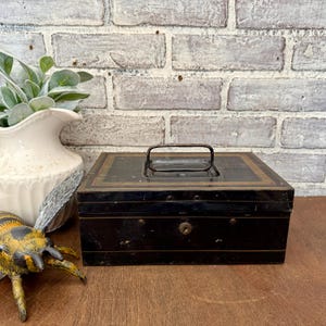 May include: A vintage black metal cash box with a handle and lock. The box has gold trim and is sitting on a wooden surface. A decorative bee and a white vase with greenery are in the background.