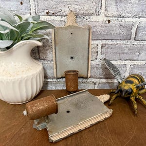 May include: A rustic, vintage-style display featuring a decorative bee figurine, two aged metal wall plaques, and a small, weathered metal container. A white ceramic pitcher with greenery adds a touch of contrast against the brick background.