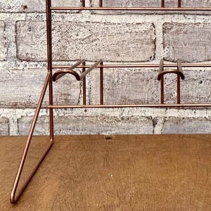 May include: A copper-colored metal display rack with multiple shelves and supports. The rack has a minimalist design with a grid-like structure. The background features a weathered brick wall and a wooden surface.