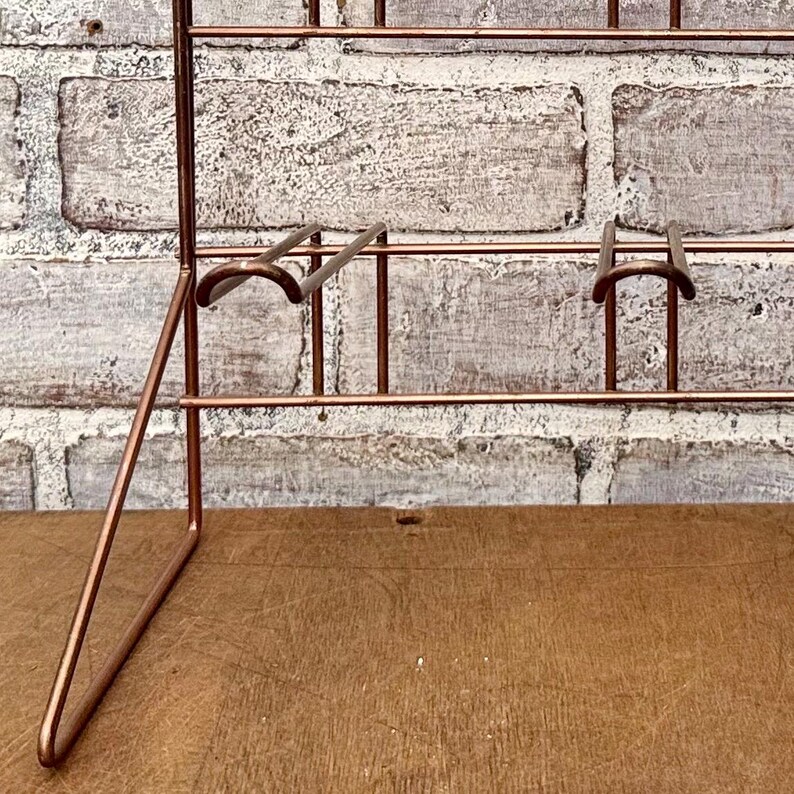 May include: A copper-colored metal display rack with multiple shelves and supports. The rack has a minimalist design with a grid-like structure. The background features a weathered brick wall and a wooden surface.