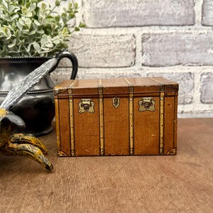 May include: A small, vintage-style brown trunk with brass hardware. The trunk has a textured surface and is accented with vertical and horizontal trim. The trunk features two brass clasps and a central lock. The trunk is a decorative storage item.