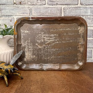 May include: A rectangular, distressed metal tray with a scalloped edge and a rusty brown finish. The tray has a worn, textured surface, suggesting age and use. Decorative items are in the background.
