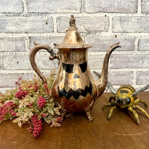 May include: A silver-colored teapot with a jack-o'-lantern face painted on the front. The teapot has a curved handle and spout, and sits on three small feet. A decorative bee and floral arrangement are next to the teapot.