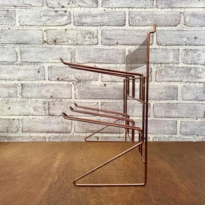 May include: A copper-colored metal rack with multiple horizontal bars for hanging items. The rack has a rectangular frame and a series of curved hooks. The background is a white brick wall and a wooden surface.