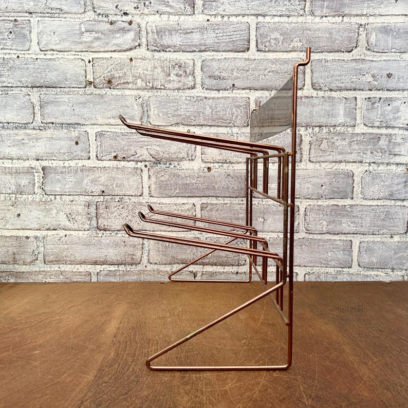 May include: A copper-colored metal rack with multiple horizontal bars for hanging items. The rack has a rectangular frame and a series of curved hooks. The background is a white brick wall and a wooden surface.