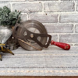 May include: A vintage metal tape measure with a red wooden handle. The tape measure is wound around a metal spool and has a metal frame.