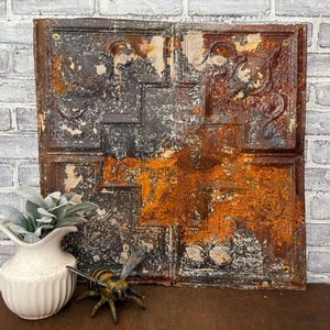 May include: A distressed metal panel with a rusty, aged appearance. The panel has a repeating square pattern with embossed floral and geometric designs. The colour palette includes shades of black, brown, and orange, with areas of peeling paint.