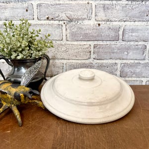 May include: A cream-coloured, ceramic lidded dish with a small, round knob on top. The dish is round with concentric rings. A decorative metal bee and a small plant in a silver container are in the background.