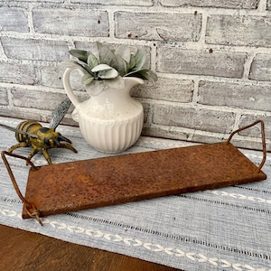 May include: A rusty metal tray with two handles. The tray is sitting on a blue and white patterned table runner. A white jug with a plant and a yellow bee figurine are in the background.