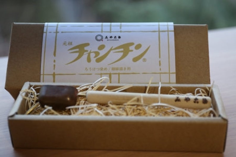 Puede incluir: Una caja de madera con una tapa marr&oacute;n y un material similar a la paja en su interior. La tapa tiene una etiqueta con el texto "元祖 チャグチ" y "ちっちゃなコップ / 携帯用コップ". Dentro de la caja hay un peque&ntilde;o objeto marr&oacute;n y un palo de madera con caracteres japoneses.