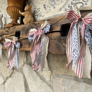 May include: A rustic wooden mantelpiece adorned with decorative bows. The bows feature red and white striped fabric, blue floral fabric, and natural linen. The arrangement creates a charming, country-style aesthetic, perfect for seasonal decor.