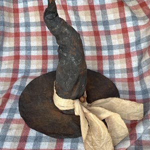 May include: A rustic, dark brown witch hat with a wide brim and a tall, slightly curved crown. A cream-colored ribbon is tied around the base of the crown. A small, metallic star adorns the tip of the hat, adding a touch of whimsy.