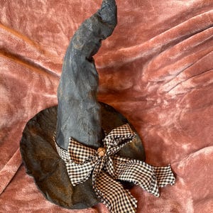 May include: A dark gray, textured witch hat with a tall, curved, and pointed crown topped with a small, rusty-colored star. The base of the hat features a black and white checkered fabric ribbon tied in a bow with a small metal bell at the center.