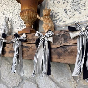 May include: A decorative garland featuring a series of fabric bows tied onto a string. Each bow is composed of multiple strips of black fabric, white fabric, and black and white striped fabric with frayed edges, accented by a small silver star.