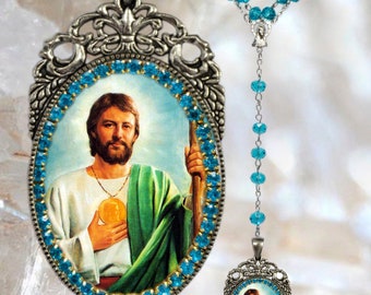 Op de afbeelding: Religieuze hangerketting met een ovale hanger met een afbeelding van een heilige, omlijst door blauwe kristallen. De ketting bevat een bijpassende rozenkransketting met blauwe kralen en een kleinere hanger. Het logo van de winkel is ook zichtbaar.