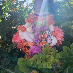 May include: A white porcelain figurine of a woman sitting cross-legged, adorned with pink, orange, and purple flowers. The figurine is holding a small framed picture of a couple, with a gold and black frame.