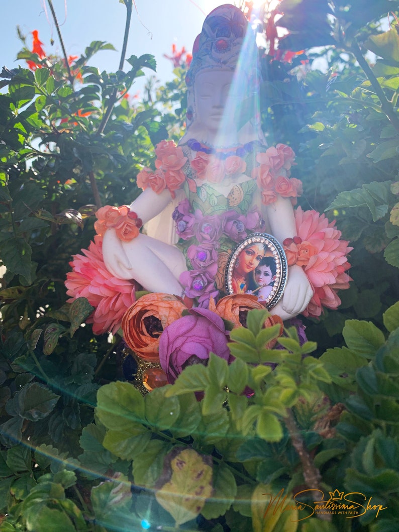 May include: A white porcelain figurine of a woman sitting cross-legged, adorned with pink, orange, and purple flowers. The figurine is holding a small framed picture of a couple, with a gold and black frame.
