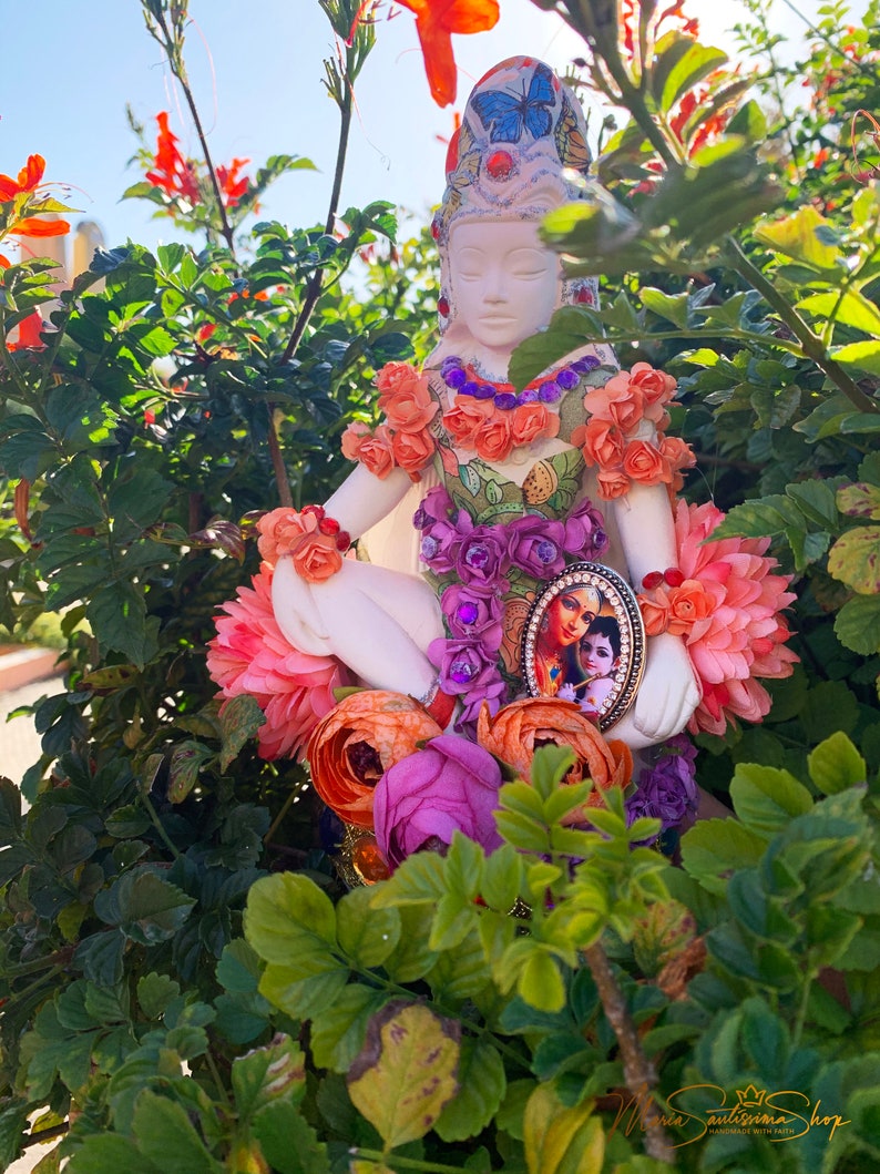 May include: A white porcelain figurine of a woman sitting in a meditative pose, adorned with colorful flowers and a butterfly. The figurine is surrounded by pink, orange, and purple flowers. A small framed image of a couple is attached to the figurine's chest.