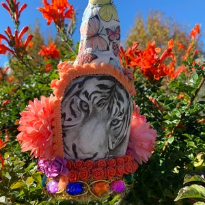 May include: A white tiger face with black stripes is painted on a ceramic bottle stopper. The bottle stopper is decorated with pink, orange, and purple flowers, butterflies, and jewels. The bottle stopper is handmade with love.