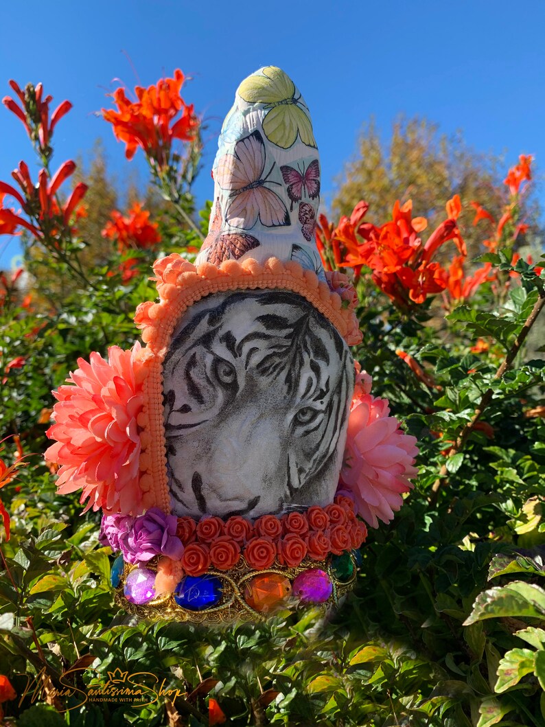 May include: A white tiger face with black stripes is painted on a ceramic bottle stopper. The bottle stopper is decorated with pink, orange, and purple flowers, butterflies, and jewels. The bottle stopper is handmade with love.