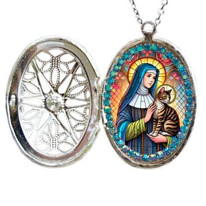 May include: Silver locket necklace with a blue and green stained glass image of a saint holding a cat. The locket is surrounded by turquoise rhinestones.