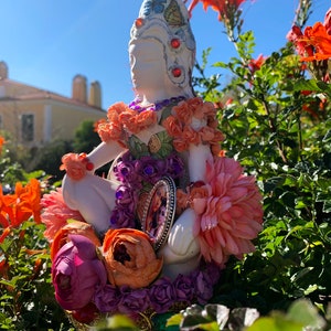 May include: A white ceramic figurine of a woman wearing a crown of flowers and butterflies, adorned with colorful beads and jewels. The figurine is surrounded by pink, orange, and purple flowers.