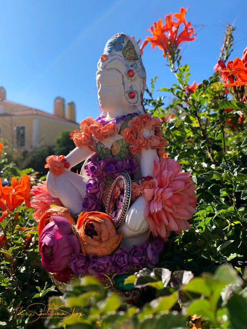 May include: A white ceramic figurine of a woman wearing a crown of flowers and butterflies, adorned with colorful beads and jewels. The figurine is surrounded by pink, orange, and purple flowers.
