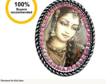 May include: An oval pendant necklace featuring a portrait of a person with dark braided hair, wearing traditional jewelry and a nose ring. The image is framed by a border of small, shimmering pink crystals and a twisted dark metal edge.