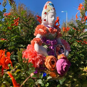 May include: A white ceramic figurine of a goddess with a colorful crown and floral embellishments. The figurine is wearing a purple and green dress with a floral pattern and is holding a small framed picture of a couple. The figurine is surrounded by orange flowers.