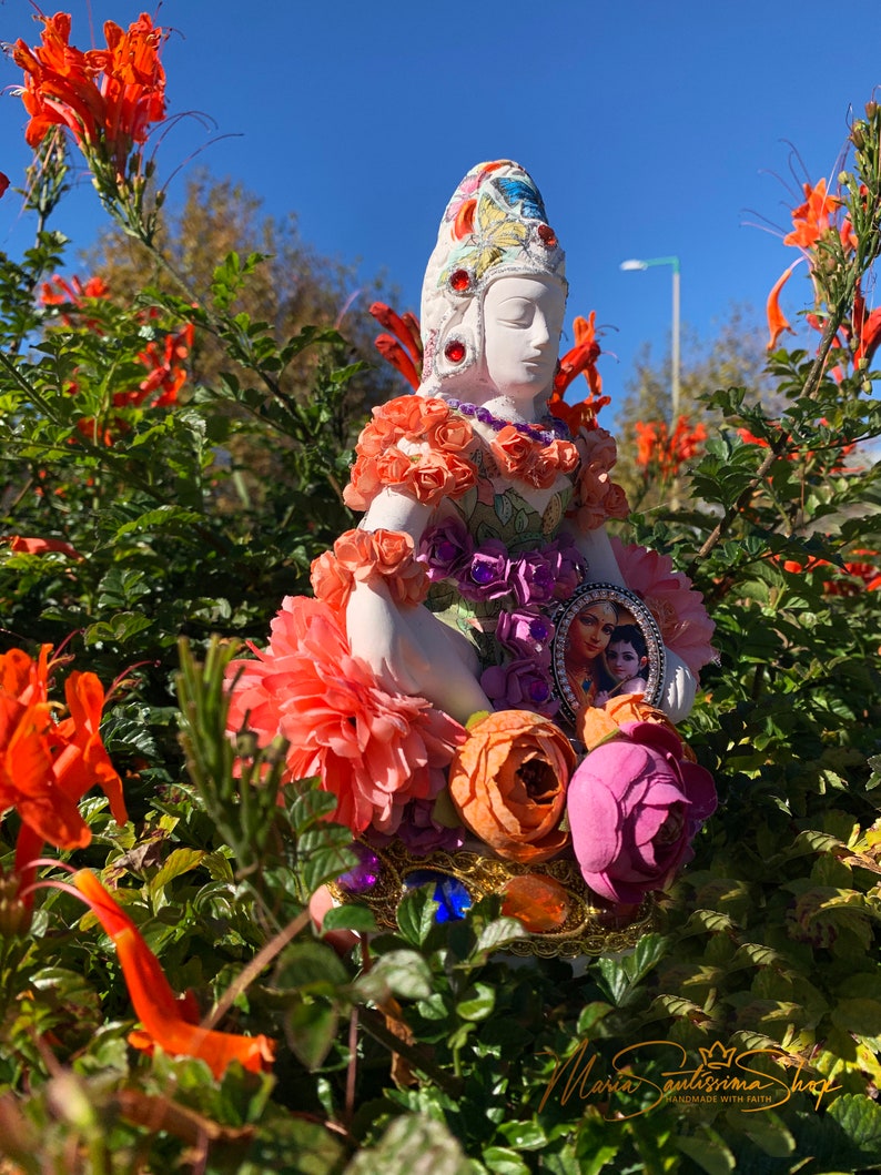 May include: A white ceramic figurine of a goddess with a colorful crown and floral embellishments. The figurine is wearing a purple and green dress with a floral pattern and is holding a small framed picture of a couple. The figurine is surrounded by orange flowers.