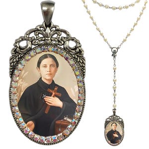 Peut inclure: Un pendentif en argent avec une image d'une femme tenant une croix. Le pendentif est entouré de strass irisés et est attaché à une chaîne en argent avec des perles blanches.