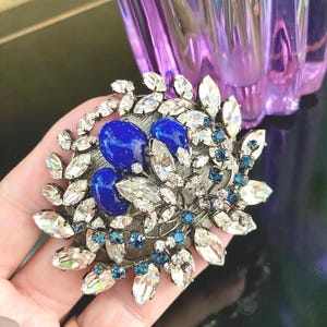 Puede incluir: Un broche de tono plateado con dos piedras azules en cabujón rodeadas de diamantes de imitación transparentes y azules.