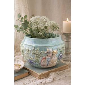 May include: A light blue ceramic planter with a detailed relief design of a couple, grapes, and foliage. The planter is filled with white flowers and greenery. A candle and a small bowl are in the background.