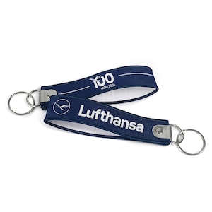 May include: Two navy blue keychains with silver key rings. One keychain has the text "100 1926|2026" in white. The other keychain has the Lufthansa logo and the word "Lufthansa" in white.