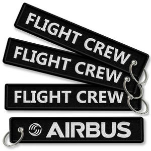 Puede incluir: Llaveros rectangulares negros con texto blanco «FLIGHT CREW» y «AIRBUS». Los llaveros tienen un anillo de metal plateado. El llavero Airbus presenta el logotipo de Airbus.