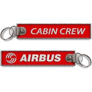 Puede incluir: Dos llaveros rojos con anillas plateadas. Uno de los llaveros dice "CABIN CREW" en blanco, el otro dice "AIRBUS" con el logotipo de Airbus. Ambos llaveros tienen un borde plateado.
