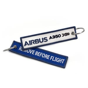 Puede incluir: Dos llaveros rectangulares de tela. Uno es blanco con "AIRBUS A350 XWB" en texto azul marino. El otro es azul con "REMOVE BEFORE FLIGHT" en texto blanco. Ambos tienen un borde azul marino y un llavero plateado.