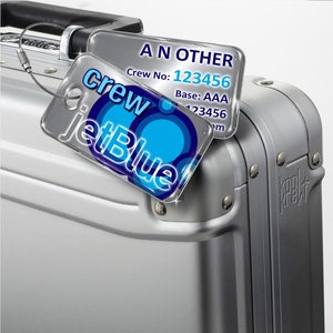May include: A silver suitcase with a blue and white JetBlue crew tag attached. The tag has the text "Crew JetBlue" and a blue circle design. The tag also has a smaller white tag attached with the text "ANOTHER Crew No: 123456 Base: AAA 123456".