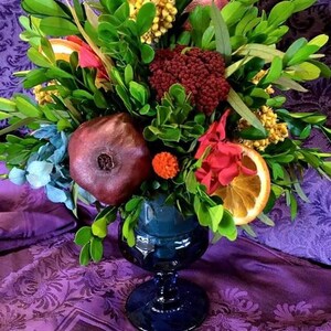 May include: A blue glass vase filled with a floral arrangement featuring a pomegranate, dried orange slices, red flowers, and green foliage.