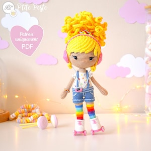 Może przedstawiać: Lalka szydełkowa z żółtymi kręconymi włosami, w różowych słuchawkach, białej koszulce z kolorowymi konfetti, niebieskim kombinezonie, tęczowymi rajstopami i białych rolkach z różowymi kółkami. Lalka stoi na białym tle z różowo-białym tłem.