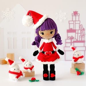 Puede incluir: Una muñeca de ganchillo con pelo morado que lleva un gorro de Papá Noel rojo y blanco, un vestido rojo con ribete blanco, un cinturón negro y botas negras. Tres pequeños muñecos de nieve de ganchillo con bufandas rojas están alrededor.