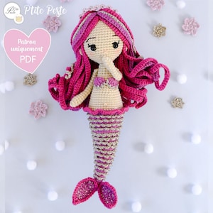 Peut inclure: Une poupée sirène en crochet réalisée avec du fil rose et or. La poupée a une queue rose, un haut rose et or et de longs cheveux roses. La poupée se tient la main à la joue.