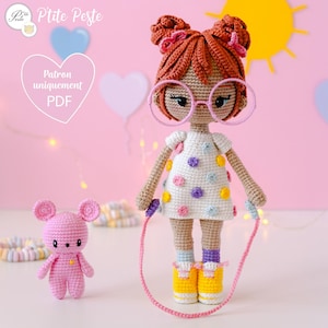 Peut inclure: Une poupée en crochet aux cheveux bruns, portant une robe blanche à pois multicolores, des lunettes roses et des chaussures jaunes. La poupée tient une corde à sauter rose. Un ours en peluche rose en crochet se tient à côté de la poupée.