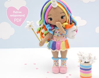 May include: A crocheted rainbow unicorn doll with a white horn, holding a white flower wand. The doll is wearing a rainbow striped dress and pink shoes. The doll is standing on a white surface with pink hearts scattered around it.
