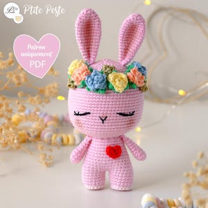 May include: A handmade pink crochet bunny with a floral crown and a red heart on its chest. The bunny has closed eyes and long ears. The image also includes text that says "P'tite Peste" and "Patron uniquement PDF".