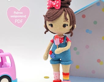 May include: A crocheted doll with brown hair, a pink bow, and a blue overall dress. The doll is holding a pink and white popsicle. The doll is standing in front of a blue and white polka dot background.